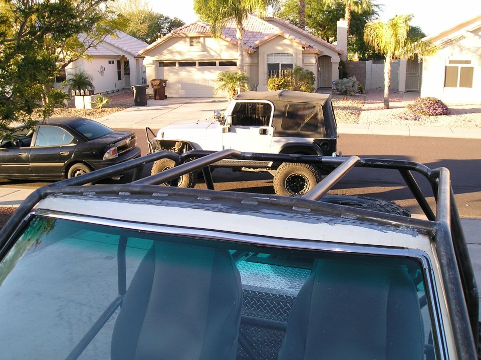 White Jeep Wrangler with soft top parked on street, black sedan next to it. Foreground shows interior of another vehicle with roll cage.
