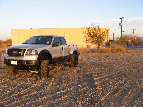 1979 Ford Bronco - A lifted, silver Ford pickup truck is parked on a gravel lot in front of a light yellow building. The truck has large, aggressive tires and…
