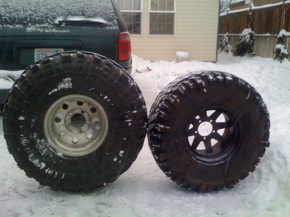 Two large off-road tires, one with a silver rim and one with a black rim, sit in the snow