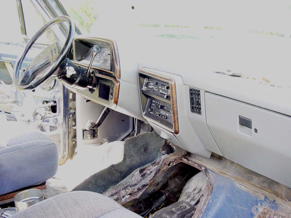 Deteriorated Ford Bronco interior with gray dash, black steering wheel, and rusted-out floor