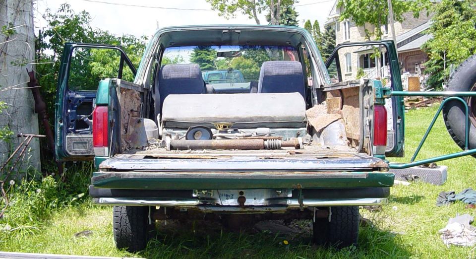 Rear view of green Bronco with tailgate down, rusty cargo bed holding driveshaft and parts, parked in grass