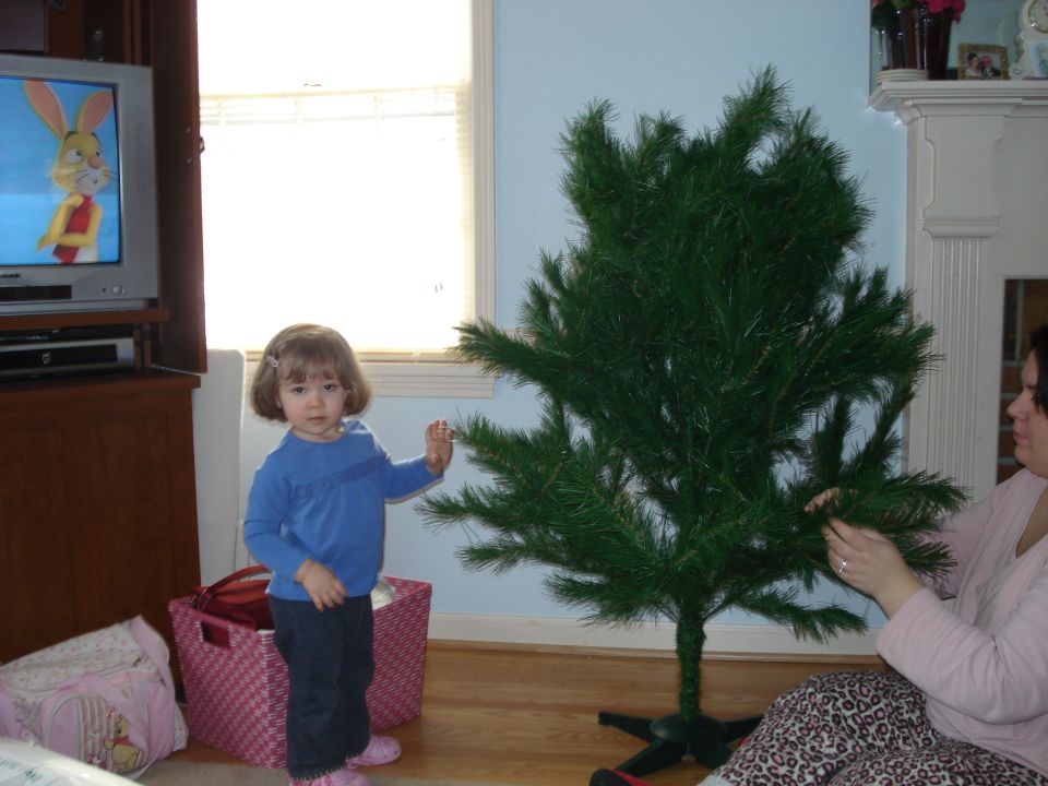 Child in blue shirt and dark pants stands by an artificial Christmas tree. A woman is assembling the tree. A TV shows a cartoon
