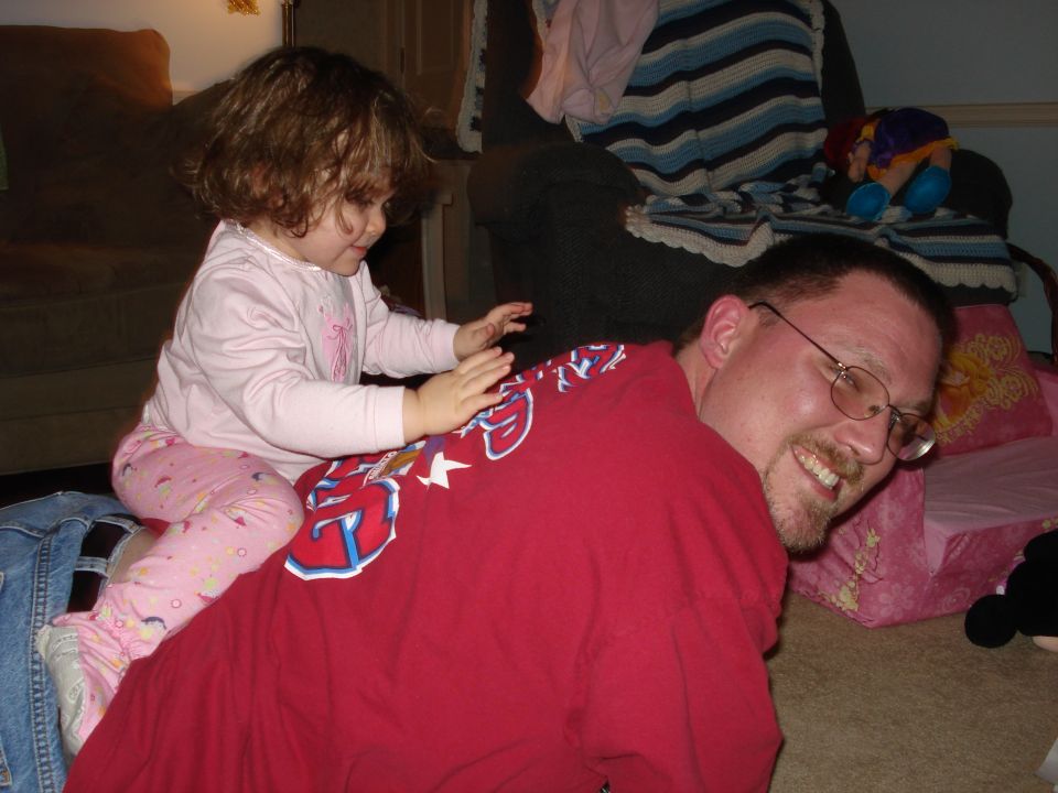 Child with curly hair sits on a man's back, both are on the floor