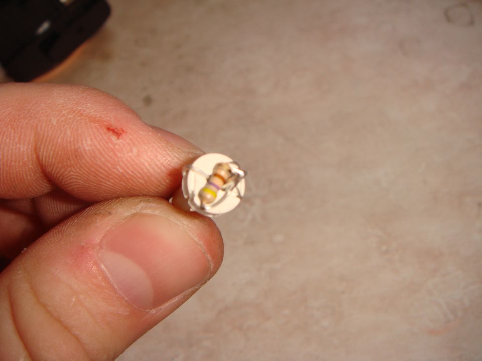 Fingers holding a tiny round circuit board with a single color-banded resistor soldered on