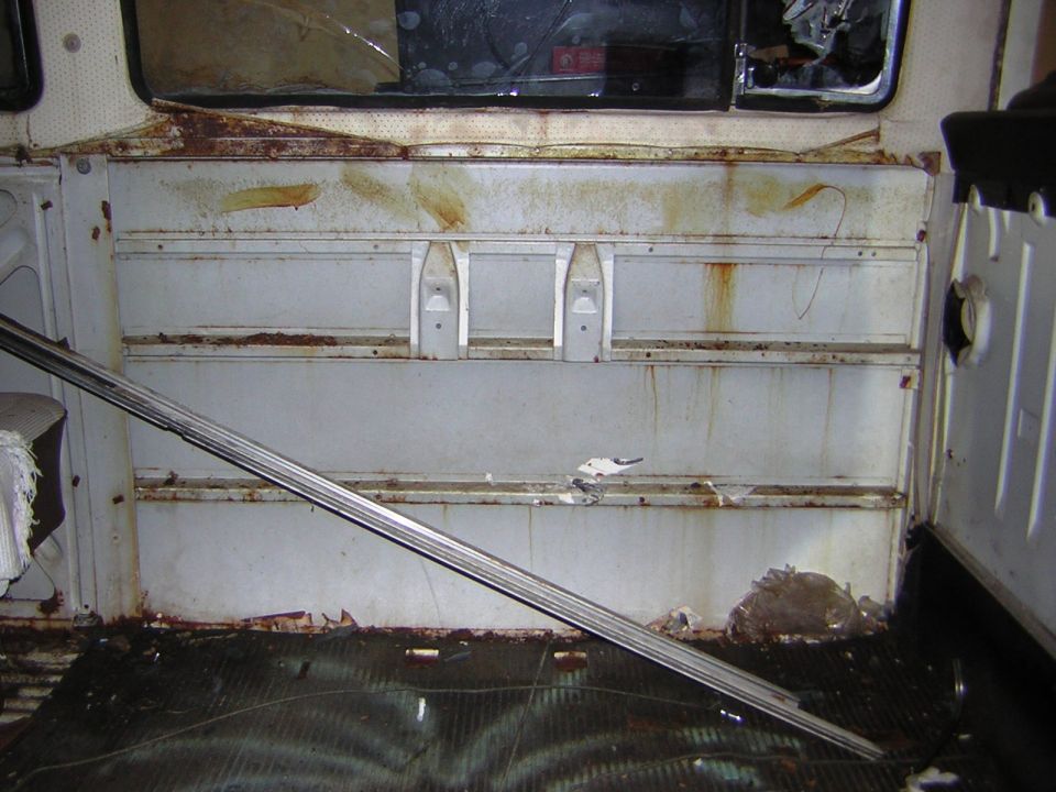 Interior of a vintage vehicle showing a rusty white wall and a silver metal bar.
