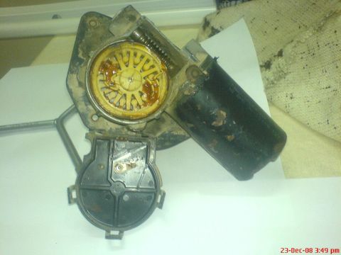 1992 Ford Crown Victoria - A wiper motor with its black plastic gear cover lifted off and set below, exposing the yellow nylon worm-drive gear swimming in old brown g…