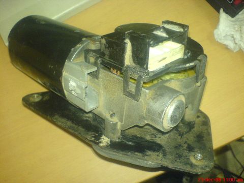 1992 Ford Crown Victoria - A wiper motor standing on its steel mounting plate atop a wood desk, showing the black motor can, cast aluminum gearbox, gray connector plu…