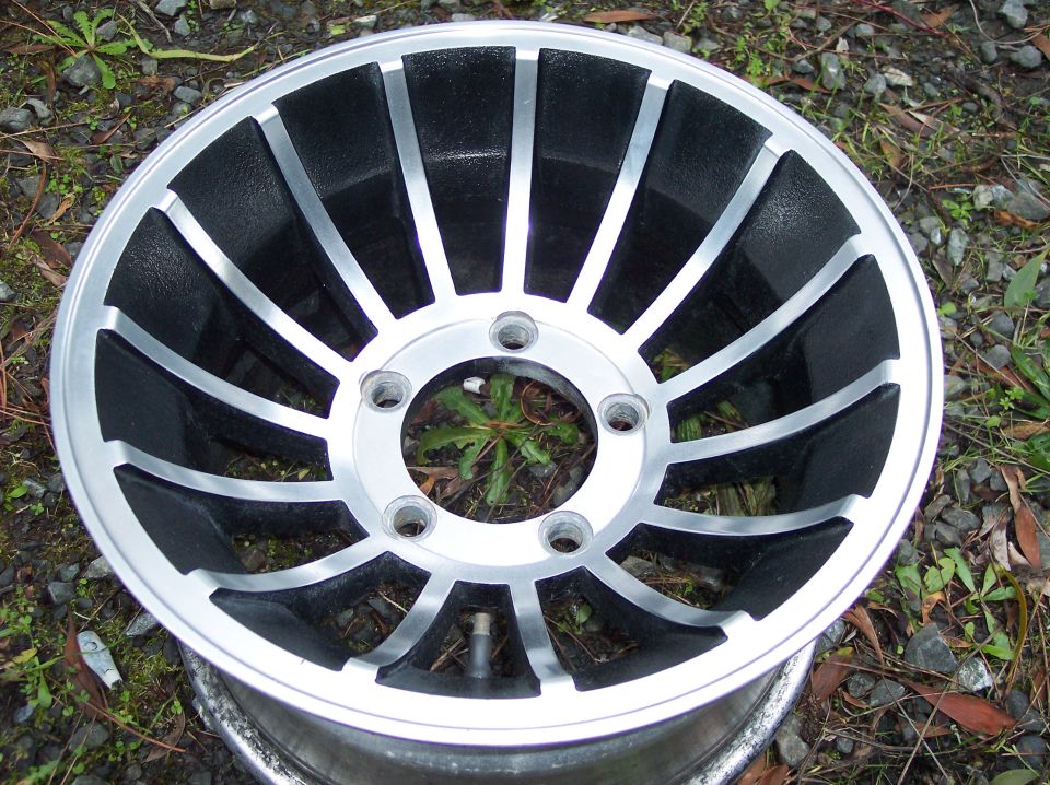 Silver and black alloy wheel with fan design, resting on gravel.