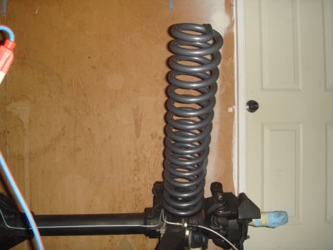 1992 Ford Bronco - A gray coil spring is mounted on a black suspension component, likely part of a vehicle's axle, against a wood-paneled wall and a white doo…