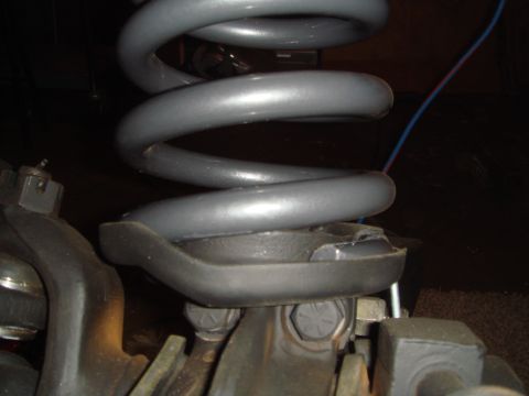 1992 Ford Bronco - A close-up view of a broken coil spring mount on a vehicle's suspension system. The gray coil spring is visible, resting on a damaged black…