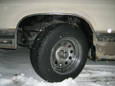 1988 Mercury Grand Marquis - A close-up side view of a vehicle's front wheel and fender well in a snowy environment. The tire is mounted on a chrome wheel with multiple…