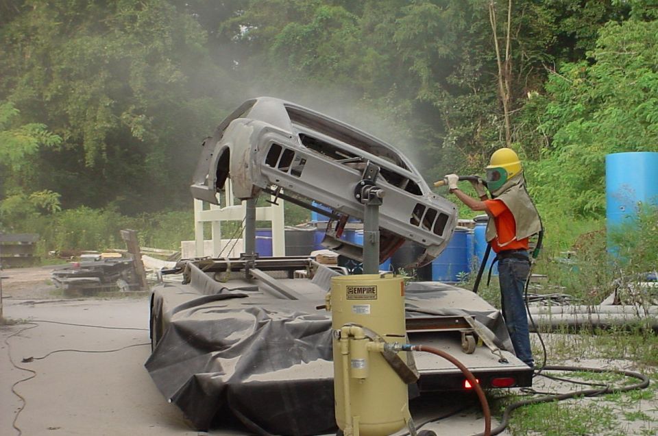 Worker media-blasting a car body shell on a rotisserie outdoors