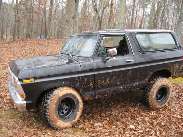 Black 1978 Ford Bronco covered in mud, parked on a leaf-covered dirt road.