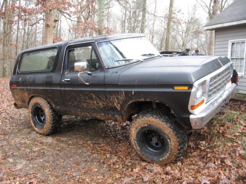 1978 Ford Bronco - A black 1978 Ford Bronco, covered in mud, sits parked on a dirt and leaf-covered ground next to a building. The vehicle has off-road tires…