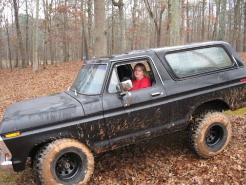 1978 Ford Bronco - A black 1978 Ford Bronco is shown from the front 3/4 angle, covered in mud, driving on a leaf-covered dirt path in a wooded area. A woman w…