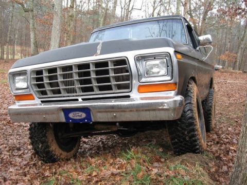 1978 Ford Bronco - Front view of a dark colored Ford truck with a chrome grille and bumper. The truck is parked on a dirt path covered in fallen leaves, with…