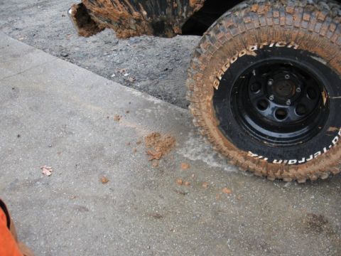 1978 Ford Bronco - A close-up view of a muddy BFGoodrich All-Terrain T/A tire on a black vehicle, with mud splattered on the concrete ground.