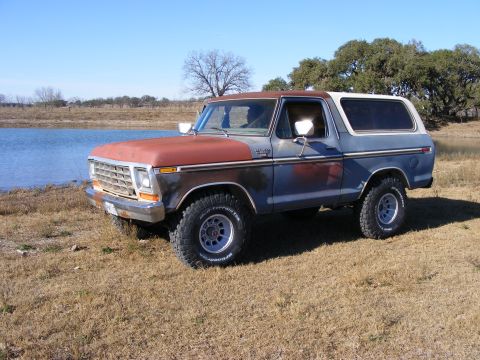 1979 Ford Bronco - A blue and rust-colored Ford Bronco is parked on dry grass next to a body of water. The vehicle has a white removable hardtop and off-road…
