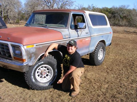 1979 Ford Bronco - A young man wearing a black t-shirt and tan pants kneels next to the front tire of a weathered, two-tone blue and rust-colored Ford Bronco.…