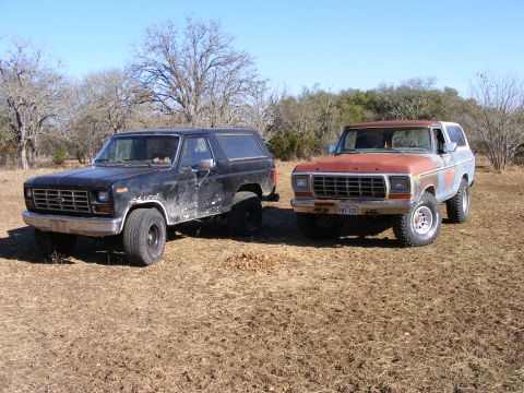1979 Ford Bronco - Two vintage Ford Broncos are parked side-by-side in a dry, grassy field under a clear blue sky. The Bronco on the left is black, while the…