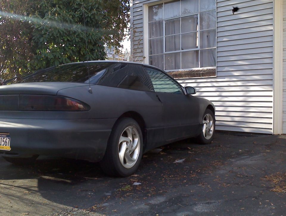 Dark gray matte finish coupe parked next to a building. Rear passenger side view.