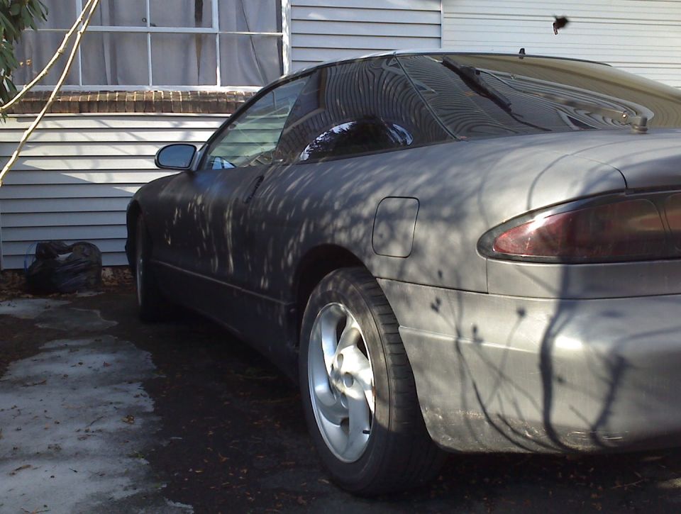 Matte gray coupe, rear passenger side view, with tree branch shadows.