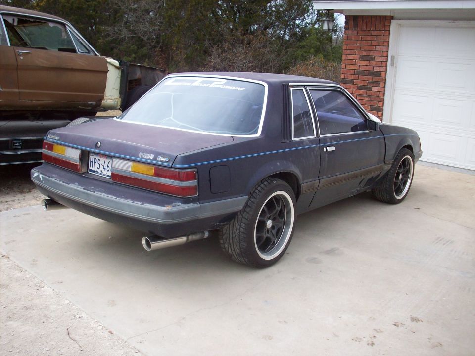 1985 Ford Mustang coupe, rear driver's side view, dark blue, black wheels, dual exhaust