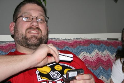 1990 Chevrolet Suburban - A man wearing glasses and a red shirt is holding two small electronic devices. He is sitting on a couch with a colorful knitted blanket.