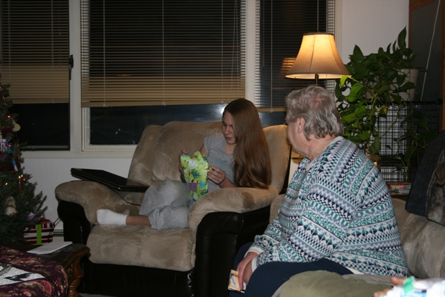Young woman opening a gift next to a Christmas tree, with an older woman watching.
