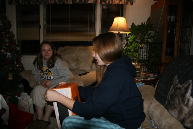 Two women in a living room, one opening a gift box, with a Christmas tree in the background.