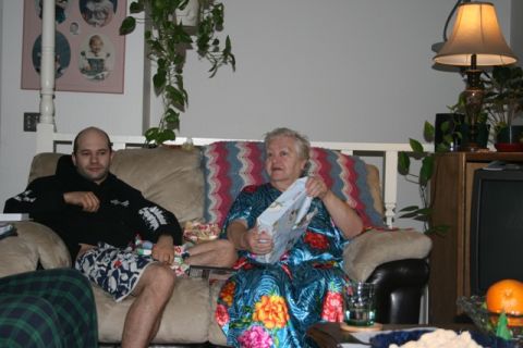 1990 Chevrolet Suburban - An older woman in a floral dress sits on a couch holding a gift box. A younger man in a black hoodie and patterned shorts sits next to her.