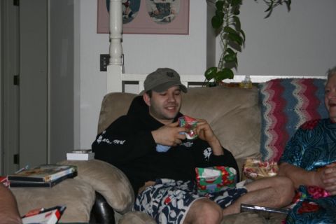 1990 Chevrolet Suburban - A man wearing a baseball cap and a black hoodie sits on a couch and holds a small gift. He is wearing patterned shorts and there are other…