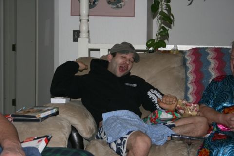 1990 Chevrolet Suburban - A man wearing a baseball cap and a black hoodie is sitting on a couch with his mouth open and his arm raised. He is wearing blue plaid shor…