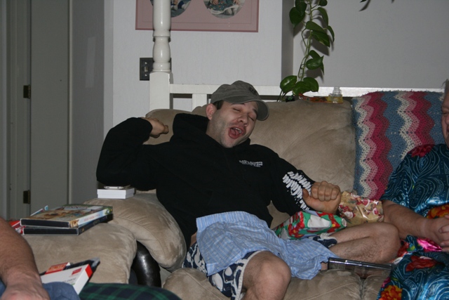 Man on couch with mouth open, wearing a hat and hoodie.