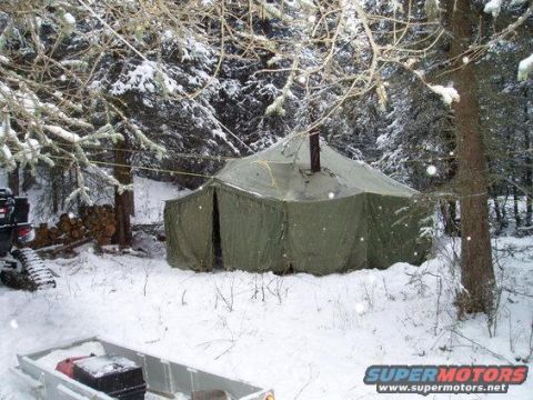 2000 Ford E350 - A green canvas tent with a chimney pipe is set up in a snowy forest. A trailer loaded with supplies and a tracked vehicle are visible in th…