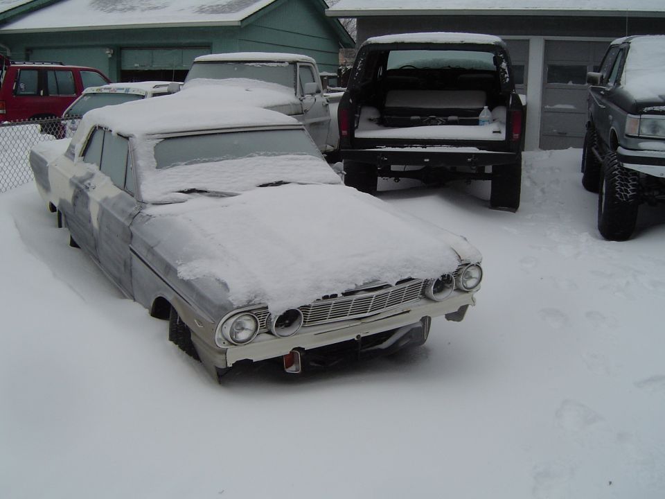 Snow-covered 1960s Ford sedan project car in primer parked in a snowy yard