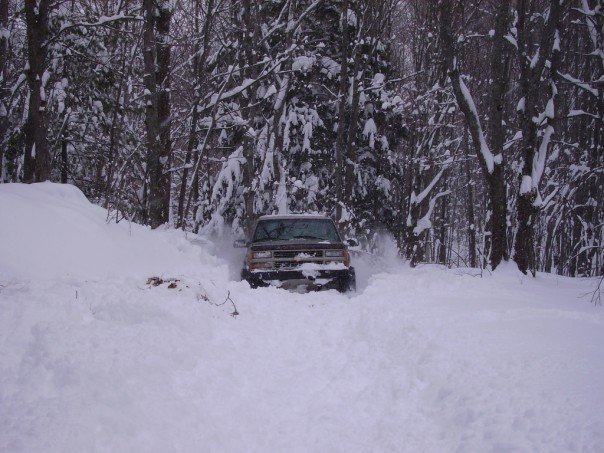 Brown Chevrolet Tahoe driving through deep snow in a forest.