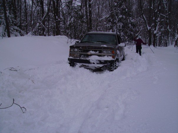Dark Chevrolet pickup truck stuck in deep snow with a skier in the background.