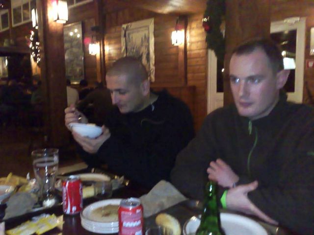 Two men sit at a table in a dimly lit restaurant, one eating from a bowl