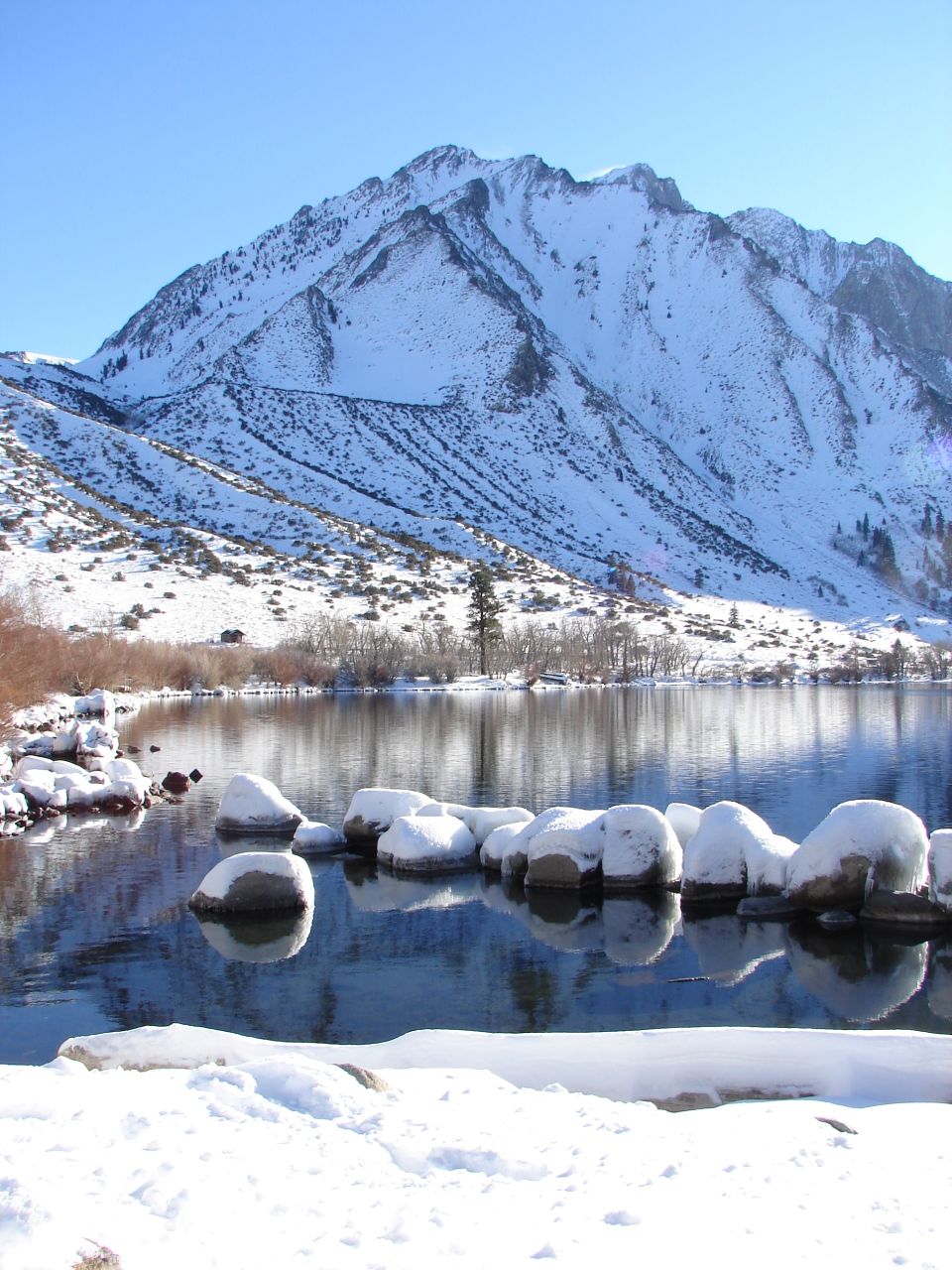 Snow-covered rocks in a lake with a snow-capped mountain and clear blue sky