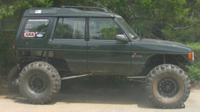 1991 Ford Bronco - Driver-side profile of a dark green Land Rover Discovery on a lifted suspension with oversized mud tires on black steel wheels and trimmed…