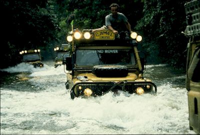 1991 Ford Bronco - Multiple Land Rover Defenders navigate a water crossing in a wooded area.