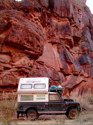 1991 Ford Bronco - Dark green long-wheelbase Land Rover Series pickup carrying a white slide-in camper, parked at the base of a towering red sandstone cliff i…