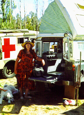 1991 Ford Bronco - A woman and a dog stand next to a customized Land Rover Defender camper, with an ambulance in the background.