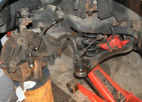 1985 Mercury Grand Marquis - Close-up of a vehicle's front suspension ball joint, which is being supported by a wooden block. A red hydraulic jack is also present in th…