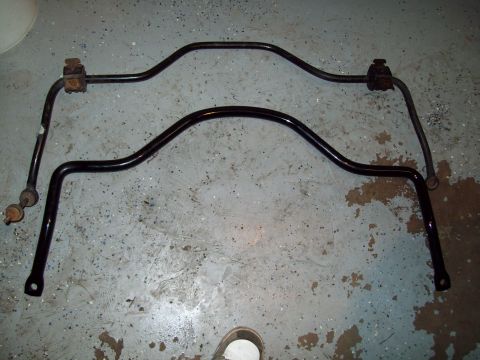1999 Mercury Grand Marquis - Two automotive sway (anti-roll) bars laid on a paint-stained concrete floor, both with black finish; the upper bar retains rusty bushing br…