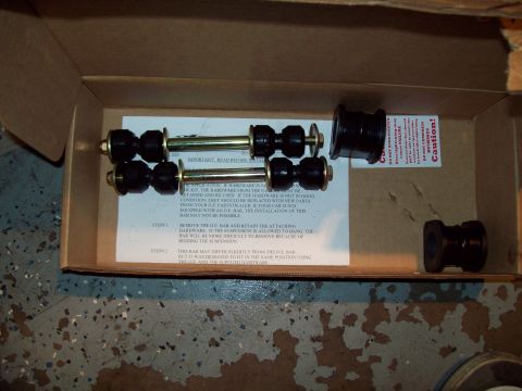 1999 Mercury Grand Marquis - Open cardboard box holding a sway bar hardware kit: a pair of gold zinc-plated end links with black rubber bushings and nuts, plus four bla…