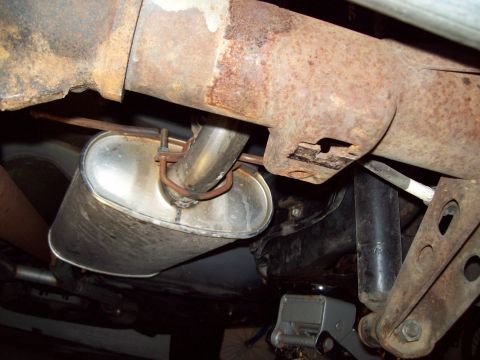 1999 Mercury Grand Marquis - Close-up underbody view of a bright aluminized oval muffler clamped with a rusty U-bolt to an exhaust pipe, tucked beneath a heavily surfac…