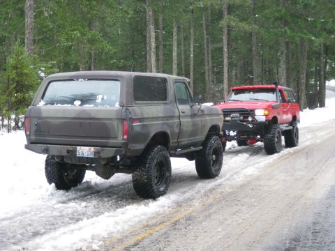 1979 Ford Bronco - Two modified SUVs navigate a snow-covered road through a forest.