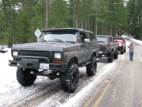 1979 Ford Bronco - A line of lifted SUVs, including a dark gray 1978 Ford Bronco, are parked on a snow-covered road in a forest.